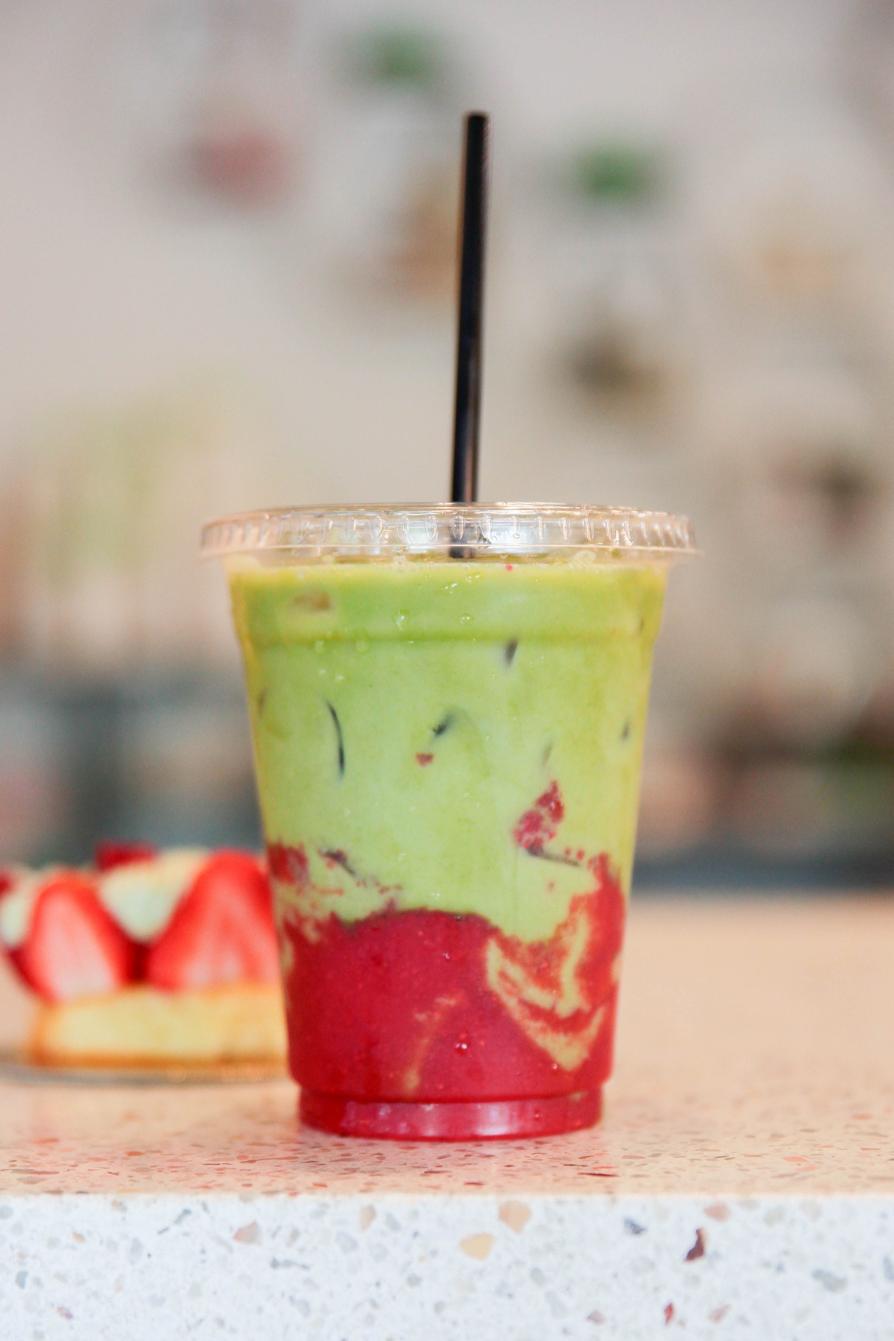 A plastic cup of iced matcha and strawberry milk, with a black straw and a pastry with strawberries in the background, all...